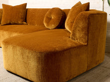 Load image into Gallery viewer, Image 13 of 16: A modern modular sectional sofa upholstered in a rich mustard-yellow velvet fabric, featuring a chaise lounge on the left and matching accent pillows, set against a white brick wall background.