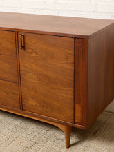 Load image into Gallery viewer, Image 12 of 24: Mid-century modern wooden sideboard cabinet with a warm medium-brown walnut finish, featuring a single drawer and two cabinet doors with dark metal pulls, set against a white brick wall and woven rug.