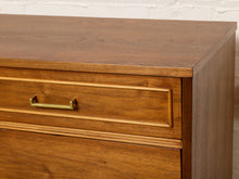 Load image into Gallery viewer, Image 12 of 21: Close-up of a mid-century modern wooden dresser with a warm medium-brown finish, featuring a single drawer with a brass pull handle and decorative trim, set against a white brick wall.
