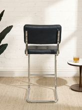 Load image into Gallery viewer, Image 5 of 17: 1970s-style counter stool featuring black button-tufted upholstered seat and backrest, polished chrome tubular cantilever frame, curved sled-style base, and integrated metal footrest.