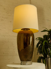 Load image into Gallery viewer, Image 8 of 17: Vintage table lamp featuring a tall rounded body with a reflective dark metallic upper finish, textured charcoal lower section, clear Lucite-style circular base, and illuminated ivory cylindrical drum shade.
