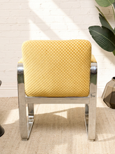 Load image into Gallery viewer, Image 5 of 15: A modern accent chair with a textured mustard yellow woven seat cushion and backrest, supported by a polished chrome metal frame, set against a white brick wall.