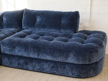Load image into Gallery viewer, Image 7 of 16: A modern sectional sofa upholstered in deep navy blue velvet, featuring a tufted button design on the seat and back cushions, set against a white brick wall background.