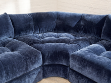 Load image into Gallery viewer, Image 6 of 16: A curved, tufted velvet sectional sofa in a deep navy blue color, shown in a close-up view against a white brick wall background.