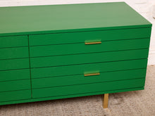 Load image into Gallery viewer, Image 8 of 15: A bright green wooden dresser with multiple drawers, featuring slim gold metal handles and a single gold leg, set against a white brick wall and textured carpet.