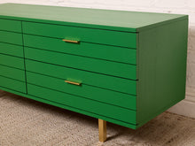 Load image into Gallery viewer, Image 9 of 15: A bright green wooden sideboard or dresser with six drawers, featuring slim gold metal handles and a single gold leg, set against a white brick wall and textured beige rug.
