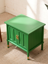 Load image into Gallery viewer, Image 9 of 21: A bright green wooden side table with a two-door cabinet design, gold-toned handles, and gold-colored caps on its legs, set on a beige carpeted floor against a white brick wall.