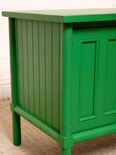 Load image into Gallery viewer, Image 11 of 21: A close-up of a bright green wooden cabinet or side table with vertical paneling on the side and paneled doors on the front, shown against a white brick wall and carpeted floor.