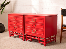 Load image into Gallery viewer, Image 2 of 21: Pair of vintage 1960s Hollywood Regency nightstands featuring vibrant poppy red painted finishes, three drawers each with brass-tone curved handles, decorative faux bamboo trim, and sculptural bamboo-inspired bases.
