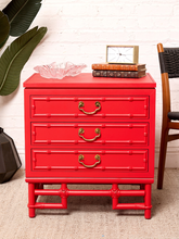 Load image into Gallery viewer, Image 4 of 21: A bright red three-drawer nightstand with a bamboo-inspired frame design, styled with a pink glass bowl, a small clock, and books on top, set against a white brick wall.
