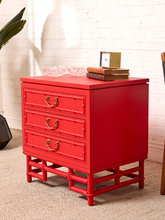 Load image into Gallery viewer, Image 5 of 21: A bright red three-drawer nightstand with decorative brass handles and a bamboo-style frame base, styled in a room setting against a white brick wall with books and a small frame on top.