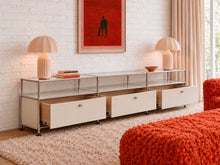 Load image into Gallery viewer, Modern three-drawer media console with chrome-toned frame, glass shelves, light neutral cabinet fronts, and open storage compartments.