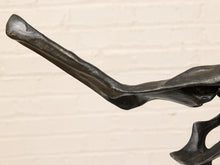 Load image into Gallery viewer, Close-up of a dark metallic sculpture or decorative object with a curved, organic shape against a light beige brick wall background.