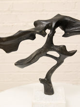 Load image into Gallery viewer, Abstract organic-shaped black sculpture on a white pedestal against a white brick wall background.