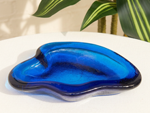 Load image into Gallery viewer, A glossy, organic-shaped blue glass dish with dark navy edges, resting on a white textured surface with a leafy plant in the background.