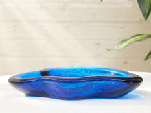 Load image into Gallery viewer, A translucent blue glass bowl with dark blue edging, displayed on a white surface against a light brick wall background with a blurred plant leaf in the corner.
