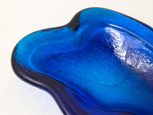 Load image into Gallery viewer, A close-up of a translucent cobalt blue glass dish with a wavy, organic shape and glossy reflective surface, photographed against a plain white background.
