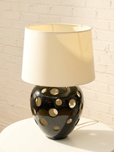 Load image into Gallery viewer, A table lamp with a cream fabric drum shade and a glossy black ceramic base featuring circular cutouts, set on a white surface against a white brick wall.