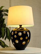 Load image into Gallery viewer, A table lamp with a black ceramic base featuring gold circular cutouts and a warm white fabric drum shade, illuminated against a white brick wall with a green plant on the left.
