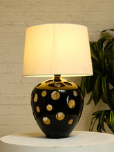 Load image into Gallery viewer, A table lamp with a black ceramic base featuring gold circular cutouts and a warm white fabric drum shade, set on a white round pedestal against a white brick wall with a green plant in the background.