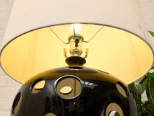 Load image into Gallery viewer, A close-up of a table lamp with a glossy black spherical base featuring circular cutouts, a warm yellow-gold interior, and a beige fabric lampshade. The light bulb is illuminated, casting a warm glow.