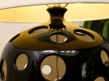 Load image into Gallery viewer, Close-up of a glossy black ceramic table lamp with circular cutouts and a warm yellow-gold interior shade, showing reflections and part of the lamp base.