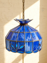 Load image into Gallery viewer, A vintage-style stained glass pendant light with a blue glass shade and bronze metal frame, hanging against a white brick wall with warm sunlight casting shadows.