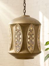Load image into Gallery viewer, A hanging pendant light with a metallic bronze finish and ornate cutout panels, shown against a white brick wall with soft sunlight and a plant leaf visible at the side.