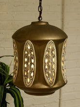 Load image into Gallery viewer, A hanging brass pendant light with ornate cutout panels that glow with warm light, shown against a white brick wall with a green plant leaf at the side.