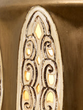 Load image into Gallery viewer, Close-up detail of a decorative carved panel with an arched, ornate cutout design in cream and dark brown tones, showing warm yellow light through the openings against a metallic gold background.