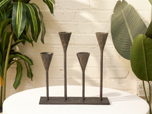 Load image into Gallery viewer, Four dark metal candle holders with a rustic bronze finish, arranged on a rectangular base on a white round table, framed by green houseplants against a white brick wall.