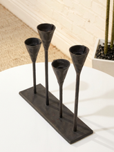 Load image into Gallery viewer, A four-arm metal candle holder with a rectangular base, featuring four tapered cup-shaped holders on slender stems, shown on a white table in a styled interior setting.