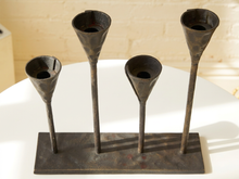 Load image into Gallery viewer, A rustic metal candelabra-style holder with four elongated cup-like candle sockets mounted on a rectangular base, displayed on a white table against a light brick wall.