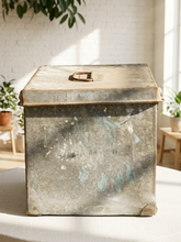 Load image into Gallery viewer, A distressed vintage galvanized metal storage chest with a hinged lid and brass handle, sitting on a white table in a bright, sunlit room with a white brick wall and plants in the background.