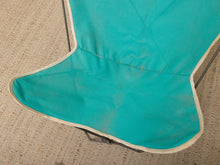 Load image into Gallery viewer, A close-up of a turquoise folding camp chair with a beige fabric trim, resting on a textured beige carpet.