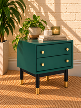 Load image into Gallery viewer, Image 2 of 5: A teal green two-drawer side table with black legs and gold-tone feet, styled with a potted plant, candle, and books against a beige brick wall.