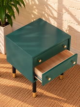 Load image into Gallery viewer, Image 5 of 5: A small side table with a dark teal green finish, featuring two drawers with brass knobs, black legs with gold caps, and a light wood interior. It is shown with one drawer open, set on a woven jute rug against a brick wall with a plant in the background.