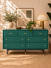 Load image into Gallery viewer, Image 1 of 4: A mid-century modern seven-drawer dresser in teal green with brass handles and legs, styled in a warm, sunlit room with a white brick wall, plants, a lamp, and a framed print.