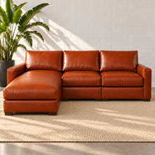 Load image into Gallery viewer, Image 2 of 14: Simulated Hollis 104-inch three-piece sectional in Mont Blanc Caramel leather with an integrated left chaise, styled on a plain ivory rug beside a bird-of-paradise plant against white brick.