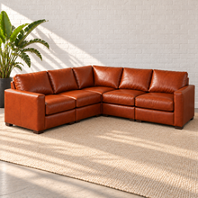 Load image into Gallery viewer, Image 3 of 14: Simulated Hollis 108-by-108-inch five-piece L-shaped sectional in Mont Blanc Caramel leather, with a square corner, styled on a plain ivory rug beside a bird-of-paradise plant.