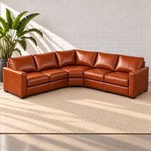Load image into Gallery viewer, Image 4 of 14: Simulated Hollis 116-by-116-inch three-piece wedge sectional in Mont Blanc Caramel leather, styled on a plain ivory rug beside a bird-of-paradise plant against white brick.