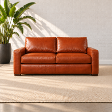 Load image into Gallery viewer, Image 3 of 3: Simulated Hollis 73-inch condo sofa in Mont Blanc Caramel leather with two seat cushions and two back cushions, styled on a plain ivory rug beside a bird-of-paradise plant against white brick.