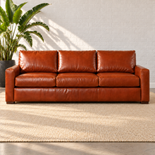 Load image into Gallery viewer, Image 1 of 3: Simulated Hollis 94-inch estate sofa in Mont Blanc Caramel leather with three seat and back cushions, styled on a plain ivory rug beside a bird-of-paradise plant against white brick.