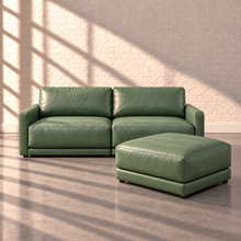 Load image into Gallery viewer, Image 2 of 3: Simulated Orinda two-piece loveseat with one separate square bumper ottoman in sage green Sequoia Rainforest leather, against white brick in natural sunlight.