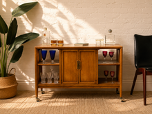 Load image into Gallery viewer, Vintage mid-century wood bar cabinet with open glassware shelves, two center doors, brass-tone hardware and caster wheels.
