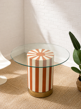 Load image into Gallery viewer, Round glass dining table with sculptural orange and cream striped cylindrical pedestal base and brass-toned trim.
