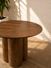 Load image into Gallery viewer, A round wooden table with a medium brown finish and a unique multi-column pedestal base, styled on a woven jute rug against a white brick wall with natural leaf shadows.
