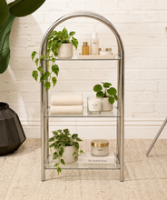 Load image into Gallery viewer, A three-tier glass shelving unit with a chrome arched frame, styled with potted plants, folded towels, skincare bottles, and candles against a white brick wall.
