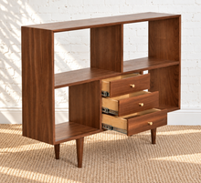 Load image into Gallery viewer, Mid-century modern wooden bookcase with open shelving and three pull-out drawers, shown in a staged room setting with a white brick wall and woven rug.
