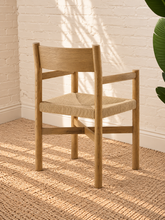 Load image into Gallery viewer, Amelie natural solid oak dining armchair with woven paper-rope seat, curved wood backrest, slim armrests, and X-shaped stretcher
